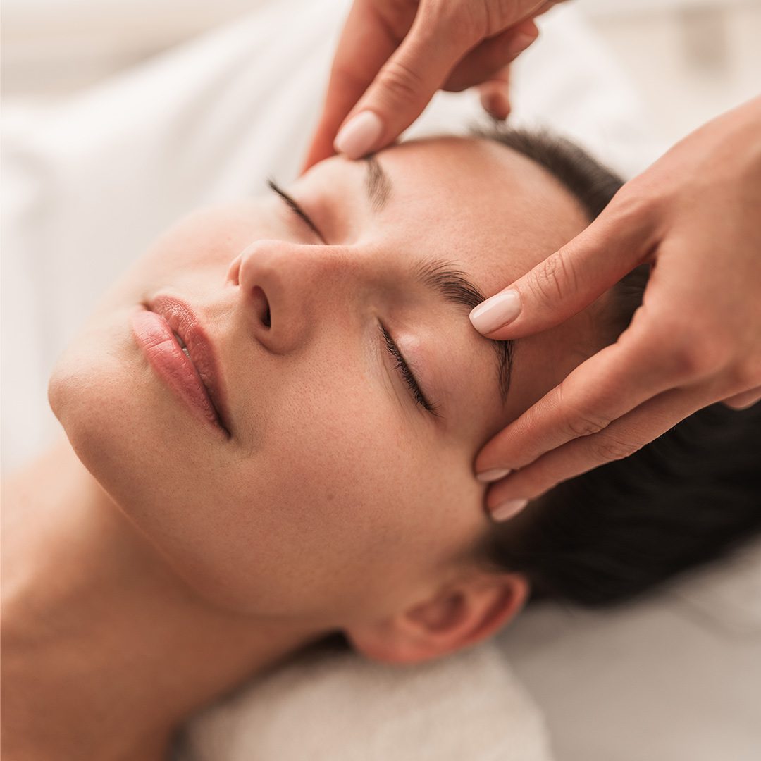 Person receiving a facial massage: practitioner gently presses temples while client rests with eyes closed in a spa setting