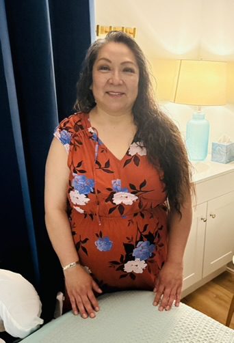 Smiling woman with long dark hair wearing a red floral dress stands beside a bed in a cozy, well-lit bedroom, hands resting on a white quilt.