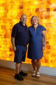 Two adults stand together and smile in front of a warm orange salt rock wall indoors, wearing casual blue clothing.