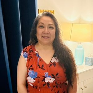Smiling woman with long dark hair in a red floral dress standing indoors by a counter with a blue lamp in the background
